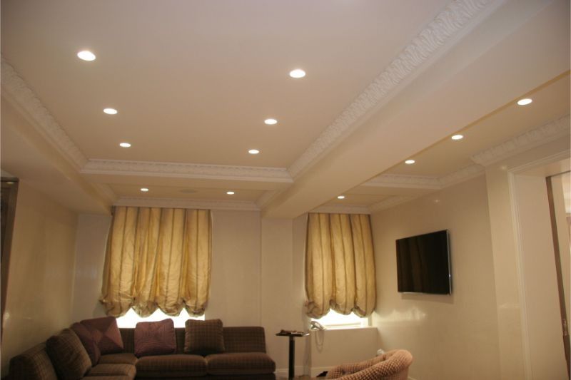 Bright living room with recessed ceiling lights, beige curtains, dark sofa, and wall-mounted TV