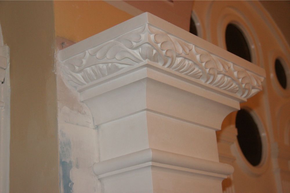 Ornate white column capital with carved leaf details against an orange interior wall