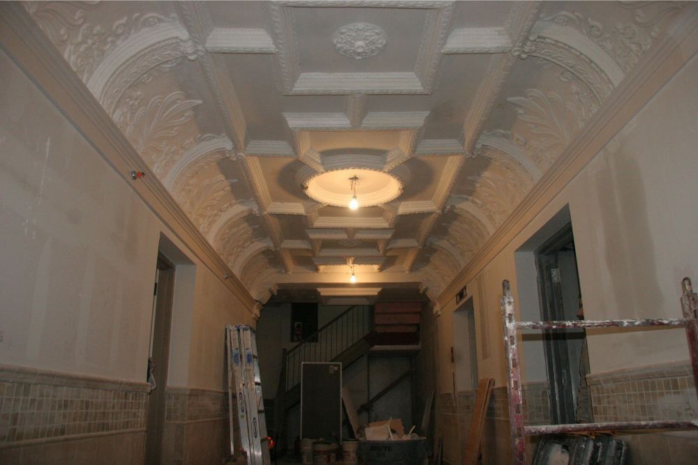 Ornate hallway with white ceiling moldings and central chandeliers, viewed from a dim interior corridor