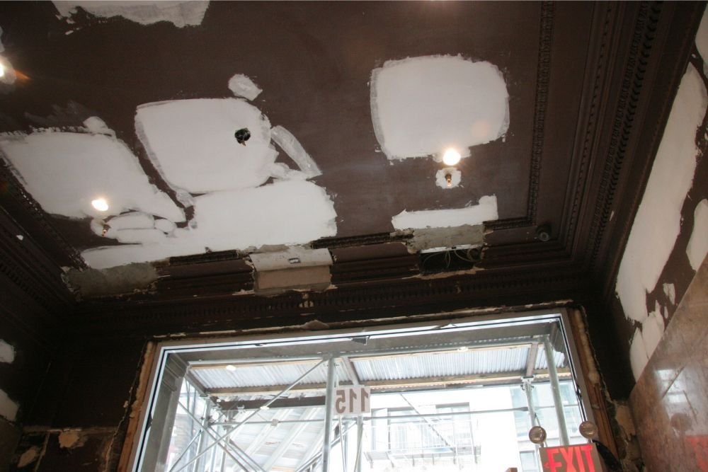 Damaged ceiling with exposed patches and lights above a doorway in a building interior