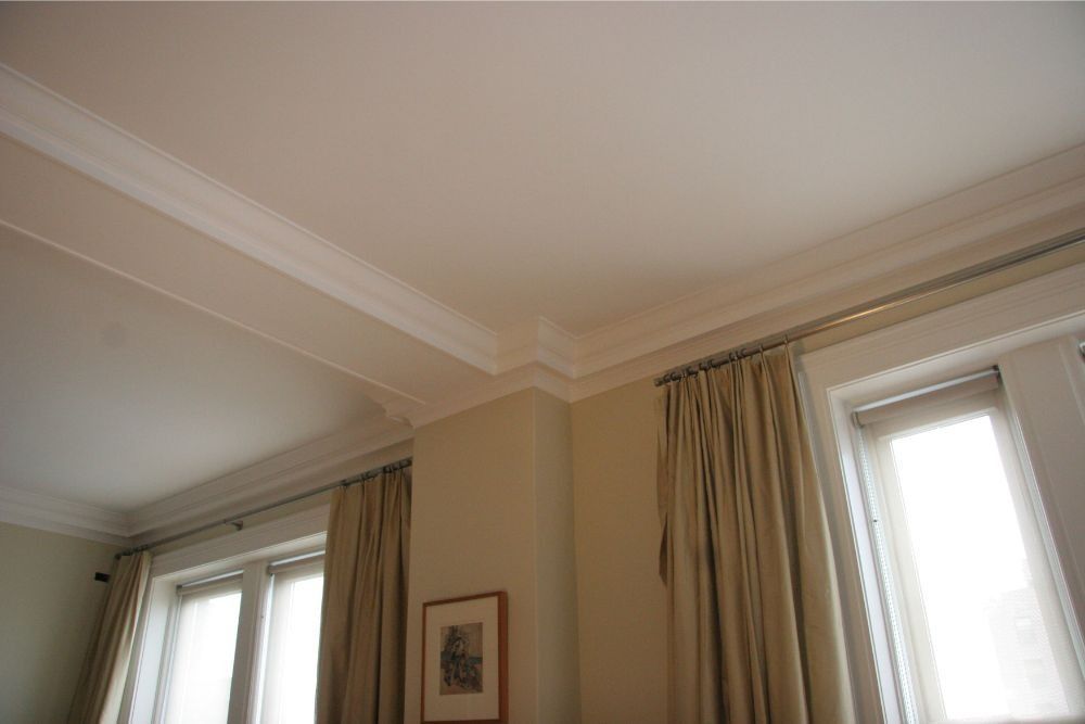 Corner of a beige room with two windows, cream curtains, and crown molding