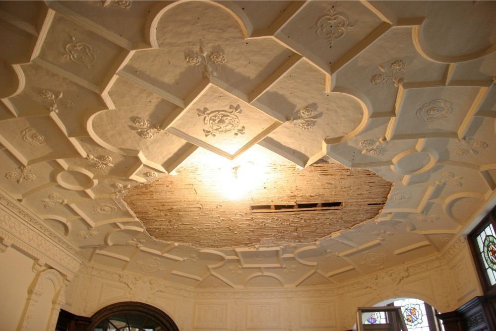 Ornate white ceiling with decorative plasterwork and a damaged patch exposing lath near a light fixture