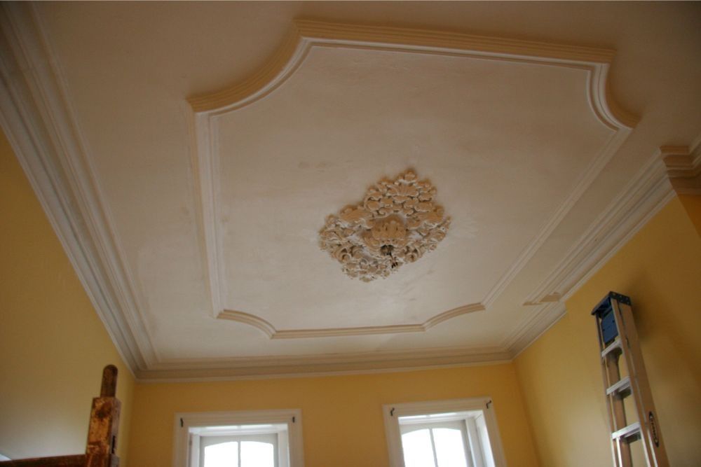 Ceiling with ornate white molding and central floral medallion above yellow walls and windows
