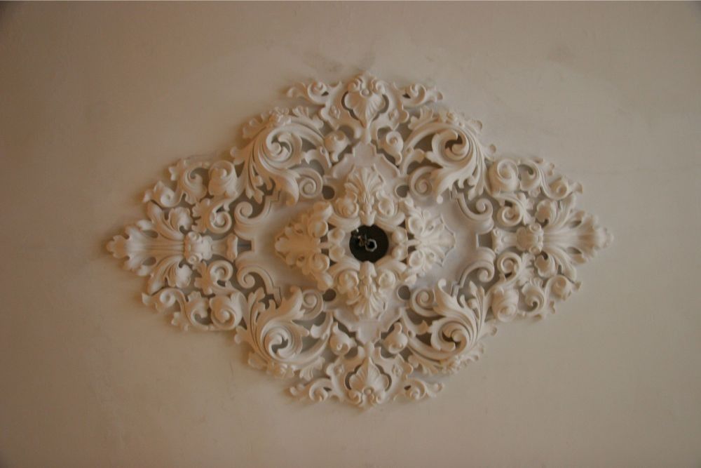 White ornate wall medallion with floral scrollwork and a dark center on a beige ceiling