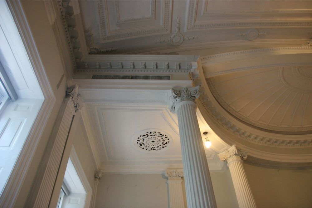 Interior ceiling with ornate white columns, moldings, and a large arched dome lit by a window.