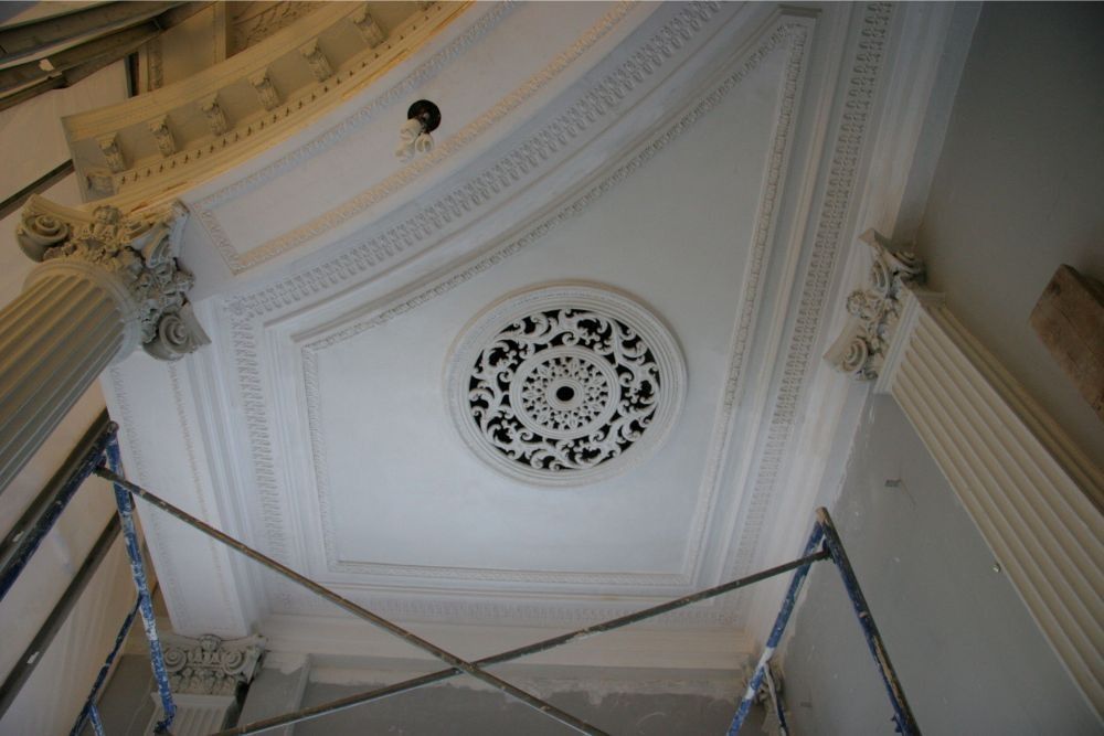 Ornate white ceiling with decorative molding and a circular vent, viewed from below with scaffolding in front