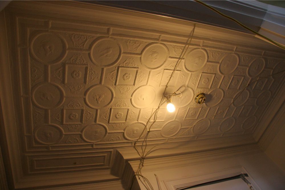 Ceiling with ornate geometric plaster design and a lit bare bulb hanging from wires