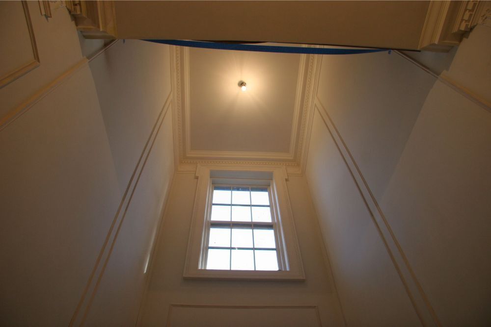 Tall stairwell with a central window and ceiling light, viewed upward between white walls