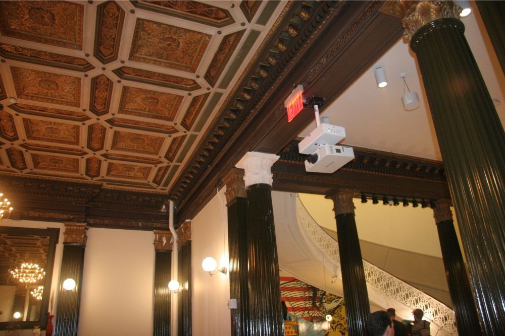 Ornate interior with carved ceiling, dark columns, wall sconces, and a staircase with a red exit sign