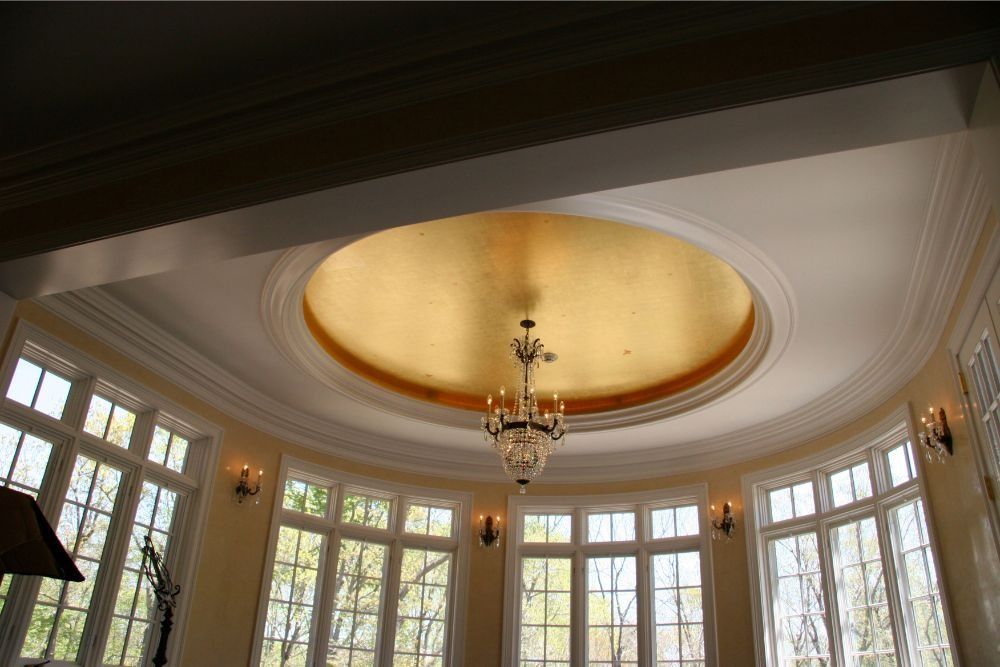 Elegant room ceiling with a gold oval medallion, chandelier, and tall bay windows letting in daylight