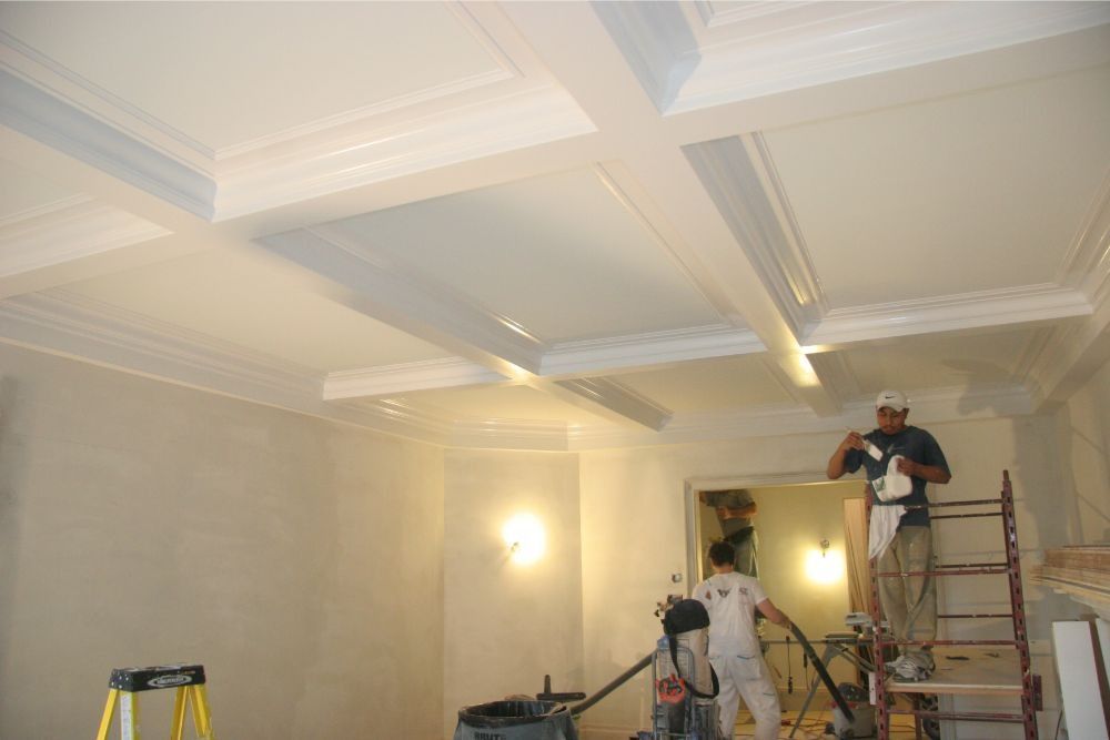 Workers painting an ornate ceiling in a room with ladders and bright lights