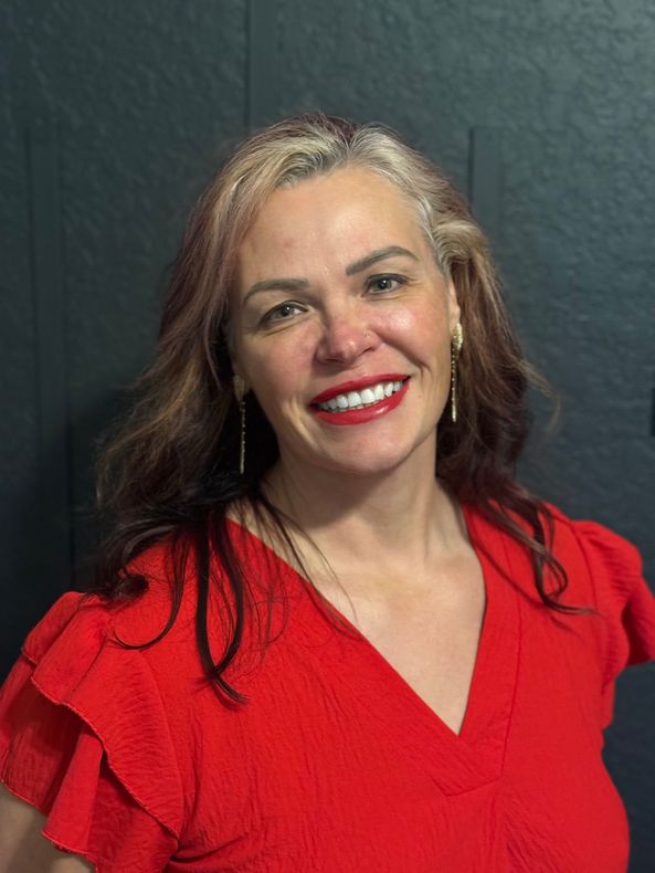 Woman with fair skin and red lipstick smiles in a red top. Against a dark wall.