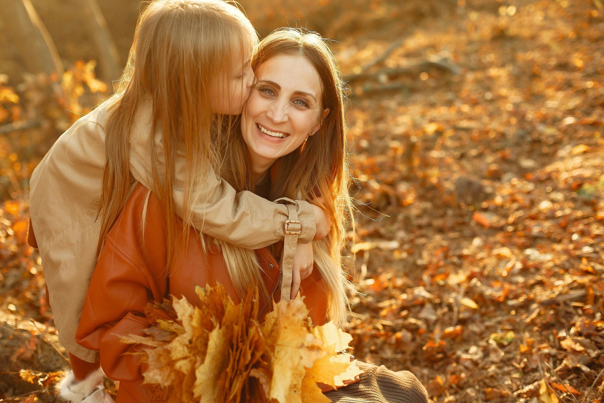 Young girl kissing smiling woman's cheek in an autumn forest; both are surrounded by fallen leaves.