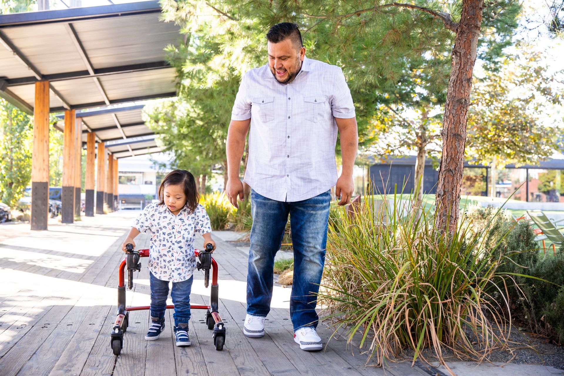 Father walking alongside a child using a walker on a wooden path. Green space with shade structure.