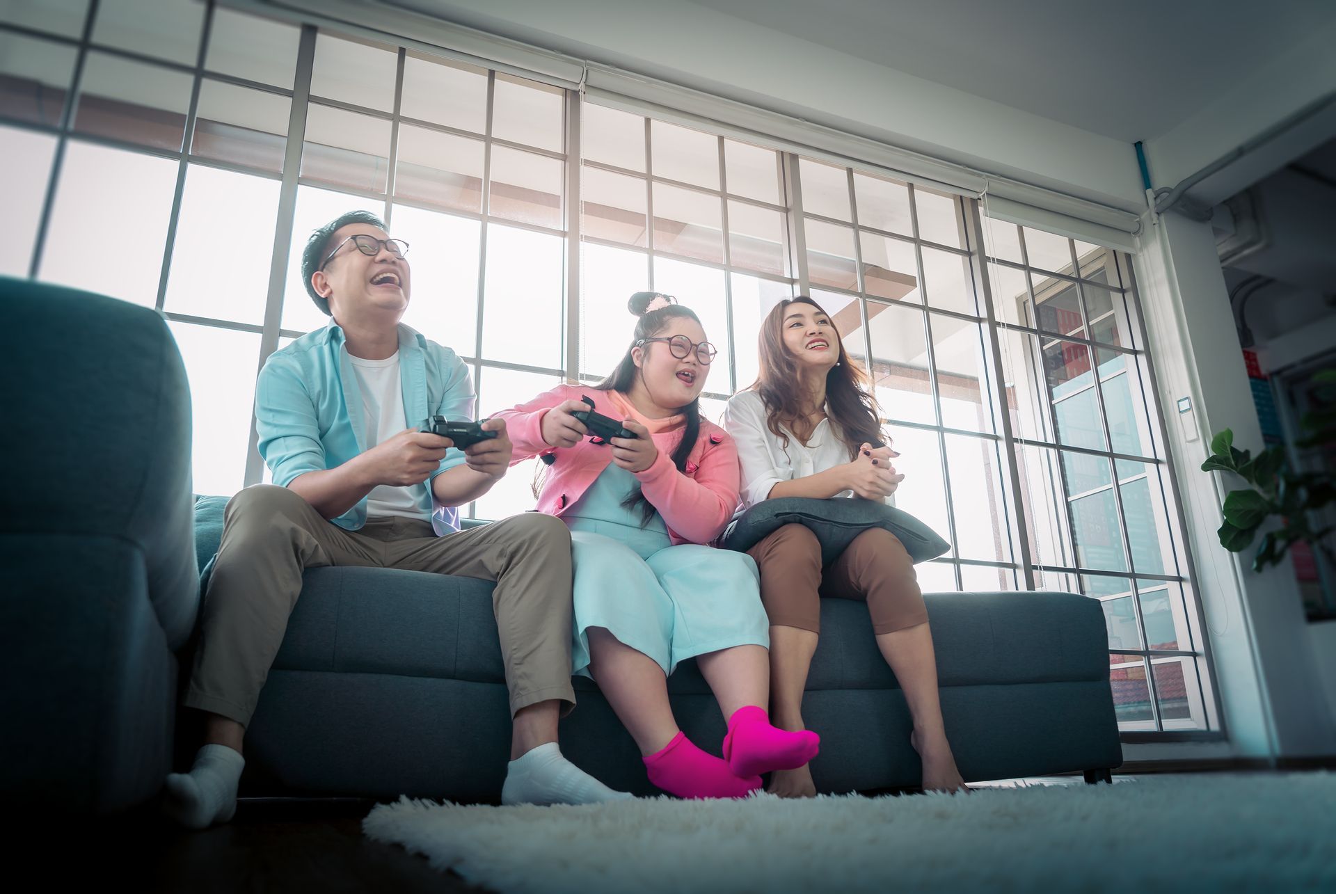 Three people laughing on a couch playing a video game in a brightly lit room.