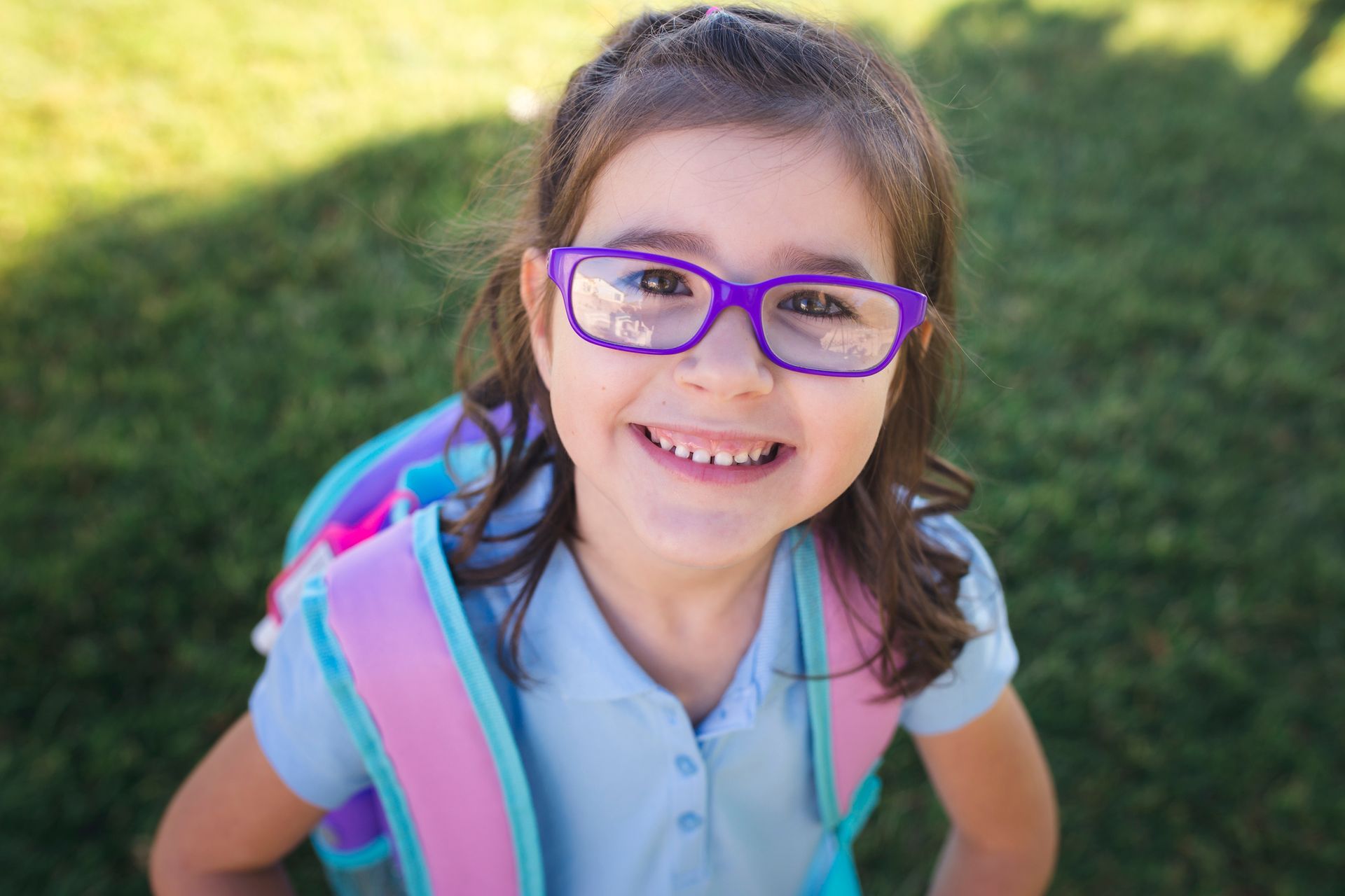 Smiling child with purple glasses and backpack stands on grass.