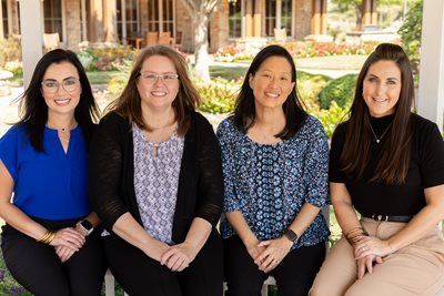 Four women seated, smiling; outdoors near a building with foliage.