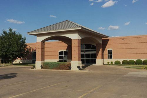 Brick building with arched entrance and glass doors under a light-colored overhang on a sunny day.