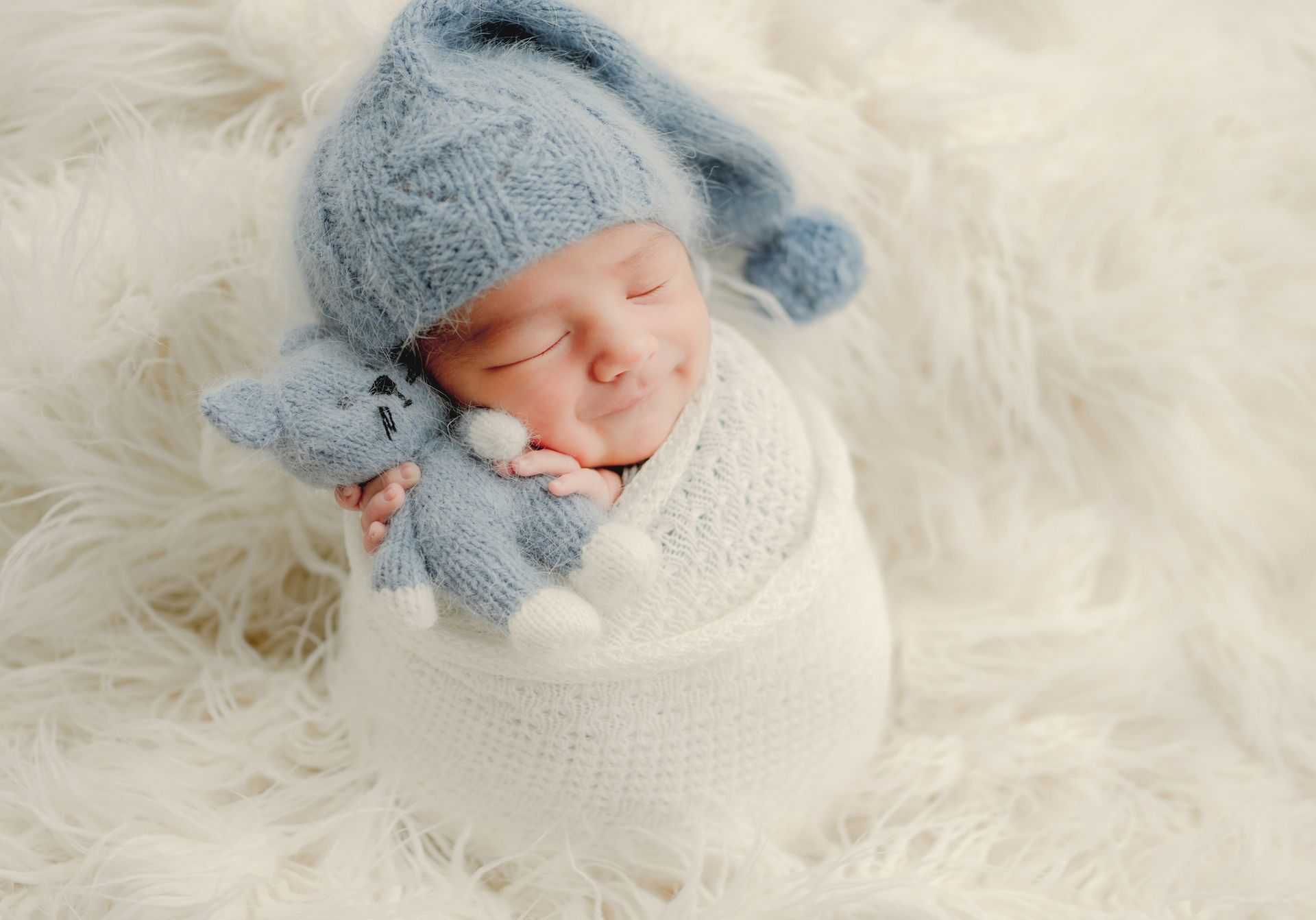 Newborn swaddled in white, wearing a blue knitted cap and holding a blue teddy bear, resting on fluffy white fabric.