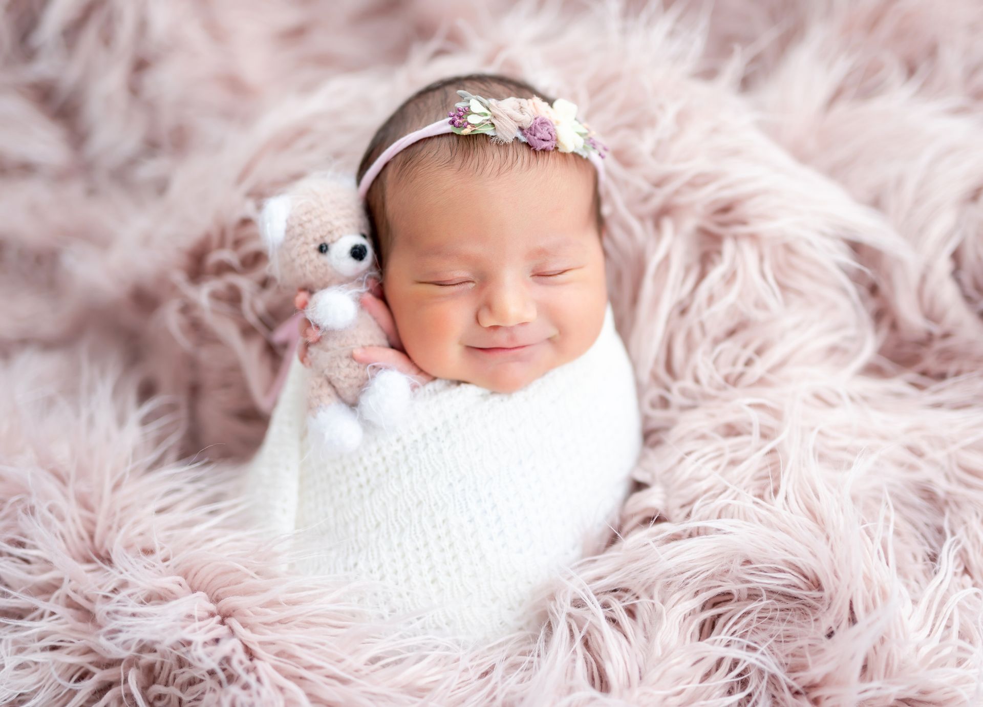Newborn wrapped in white blanket, smiling with a small teddy bear, on pink faux fur, floral headband.