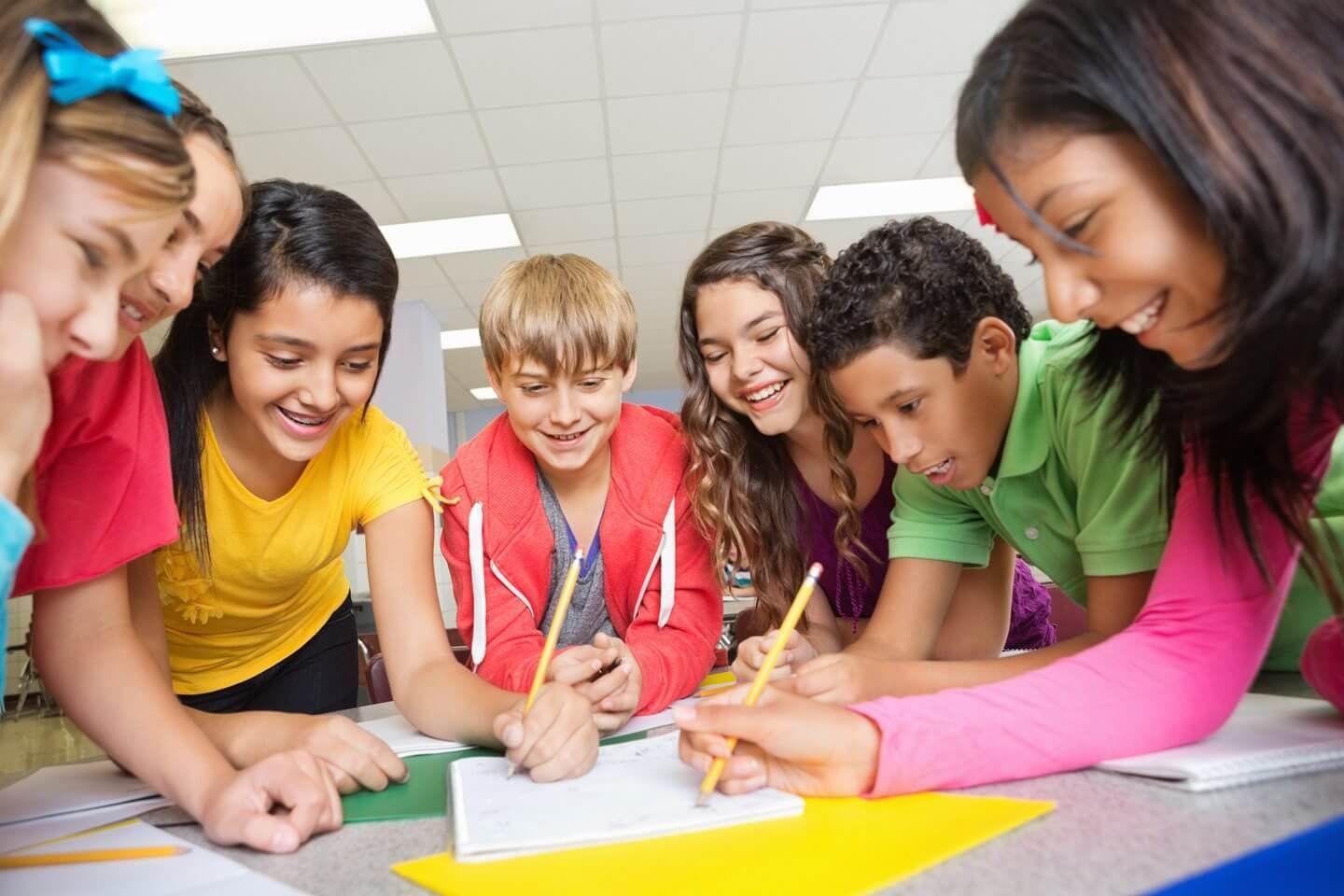 Group of students smiling and writing together at a table. Pencils, paper, and colorful clothing present.
