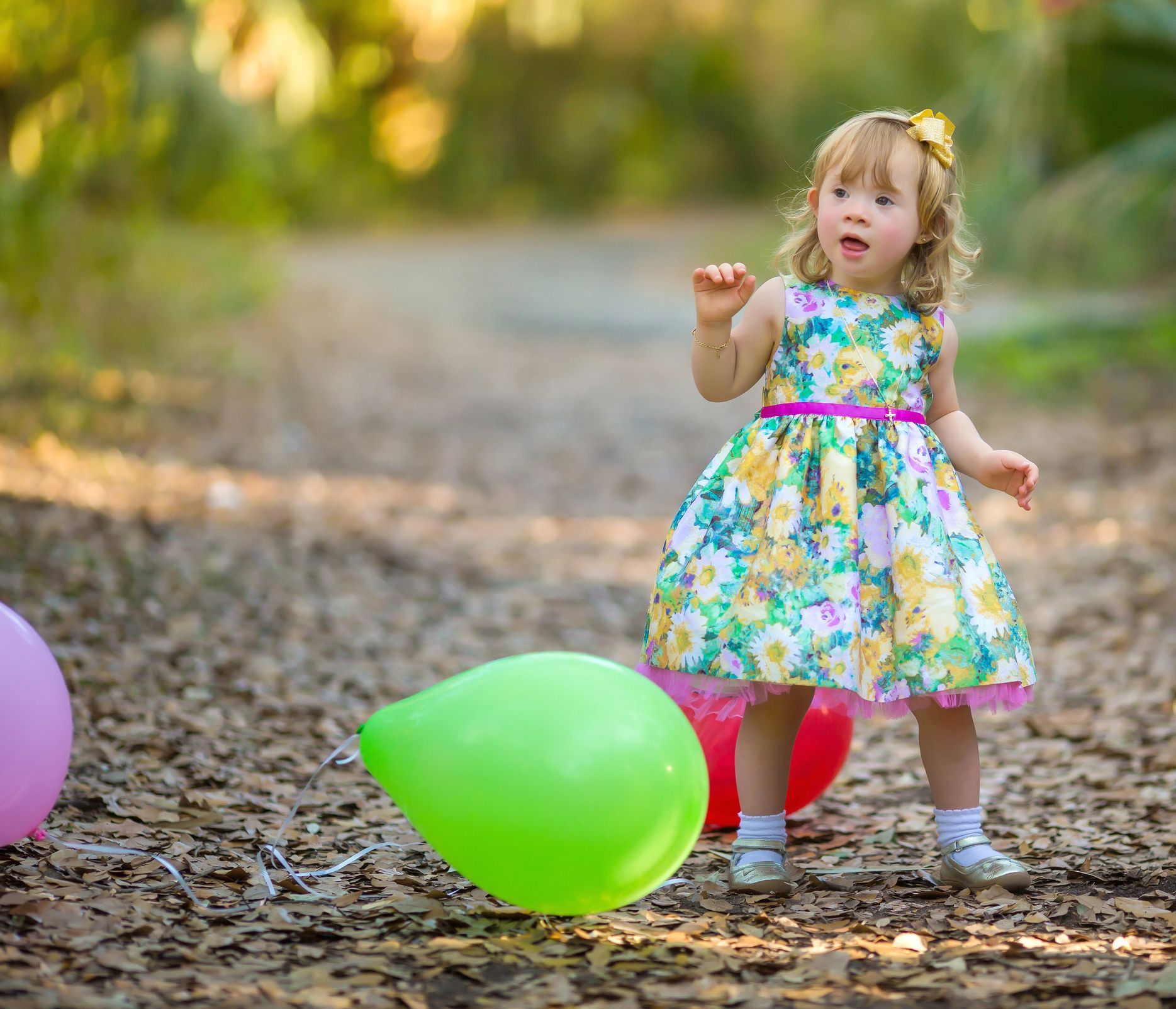 Girl in floral dress with balloons on a path. She has curly hair and a joyful expression, raising her hands.