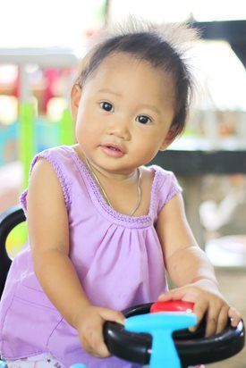 Child in a lilac dress holding a toy steering wheel, smiling at the camera.