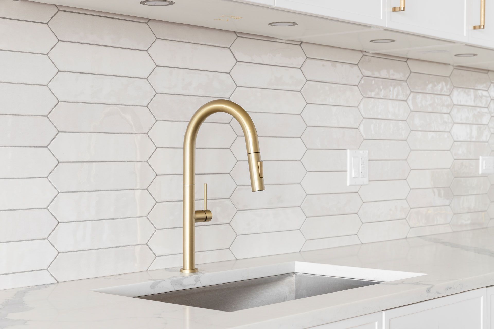 Gold faucet over a white kitchen sink with white and gray backsplash tiles.