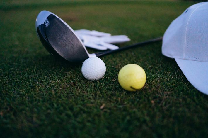 Golf clubs, a white ball, and a yellow ball on green grass beside a white cap