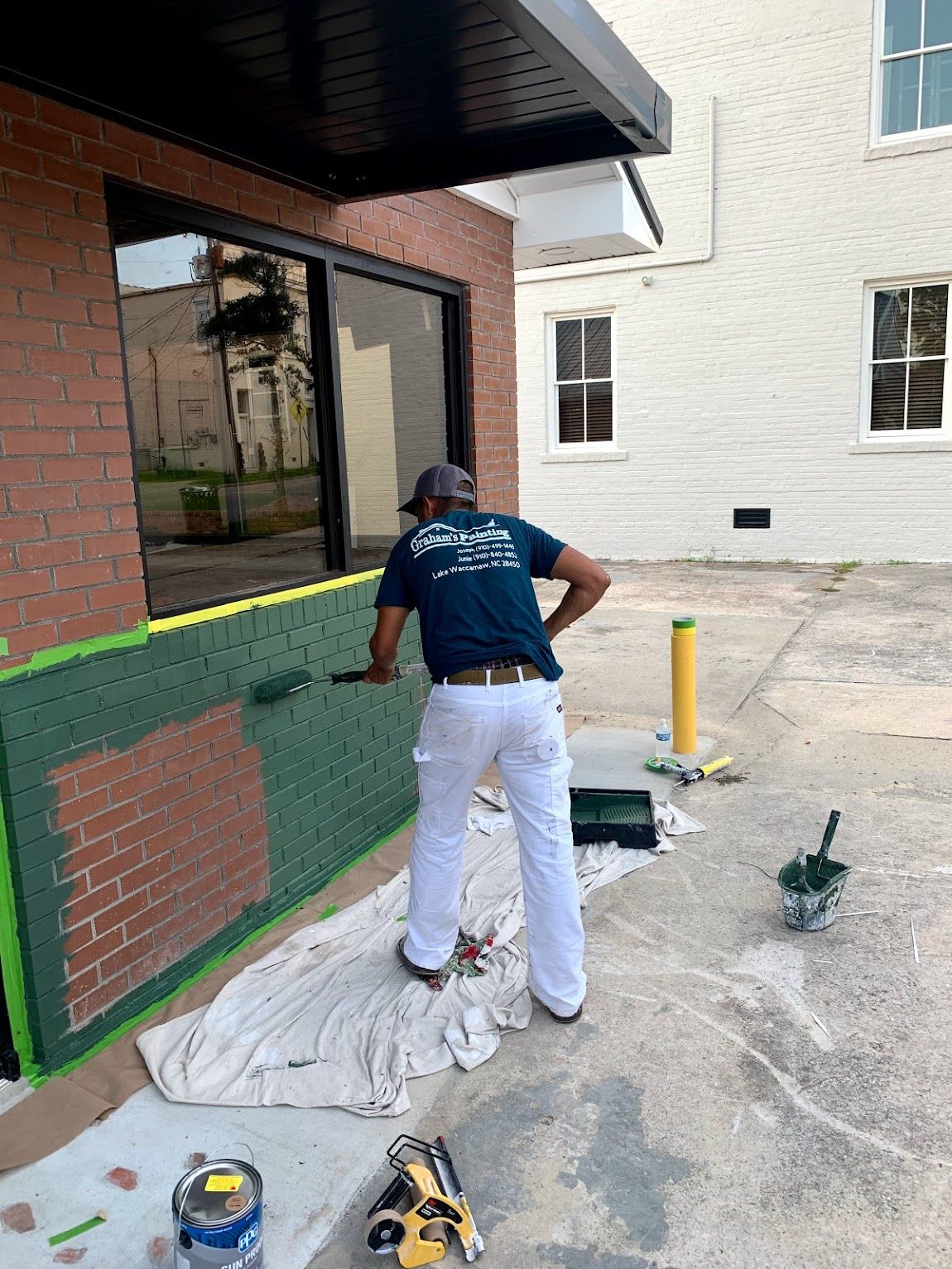 Painter in white pants and blue shirt rolls green paint on a brick wall.