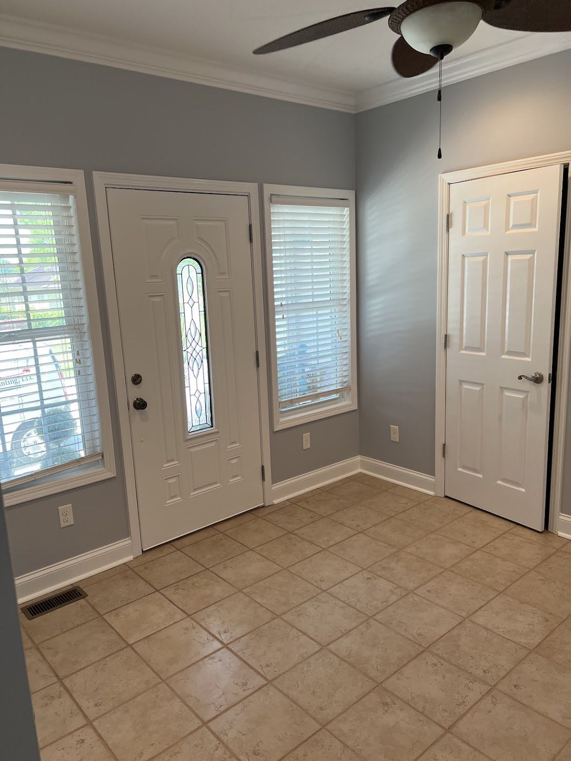 An empty room with a front door, windows, and a white interior door. Gray walls, beige tile floor.