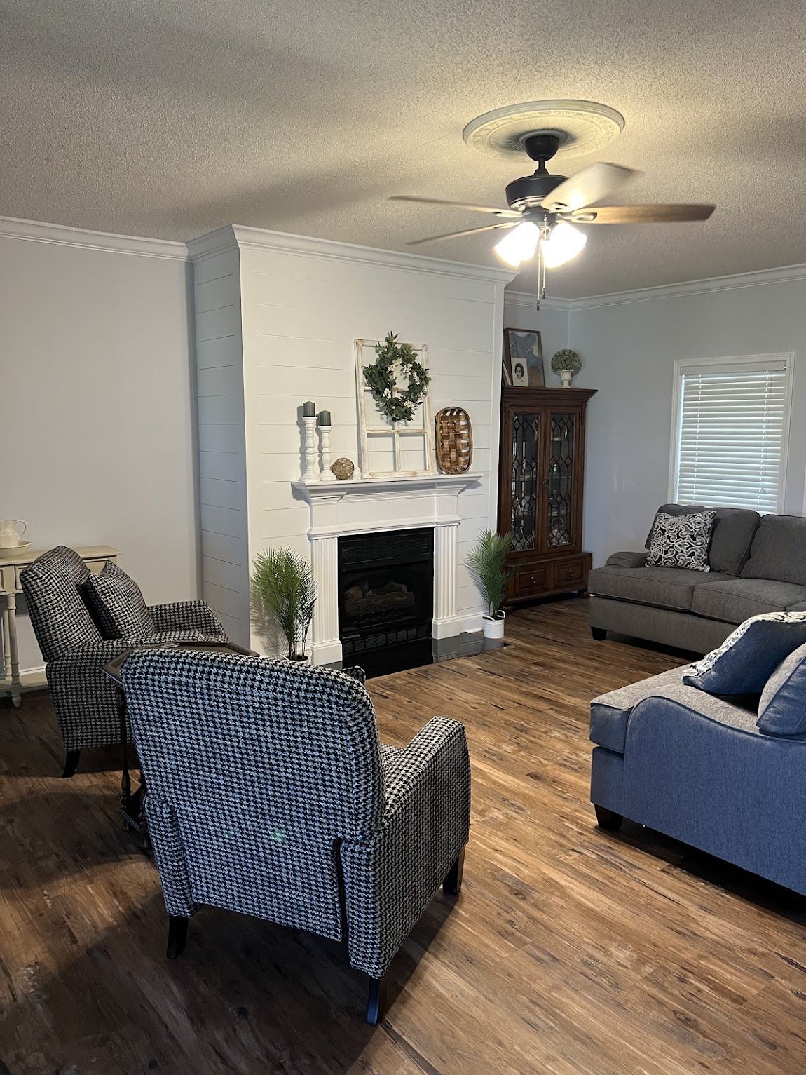 Living room with fireplace, armchairs, sofa, and wood flooring. Neutral-toned walls and ceiling fan.