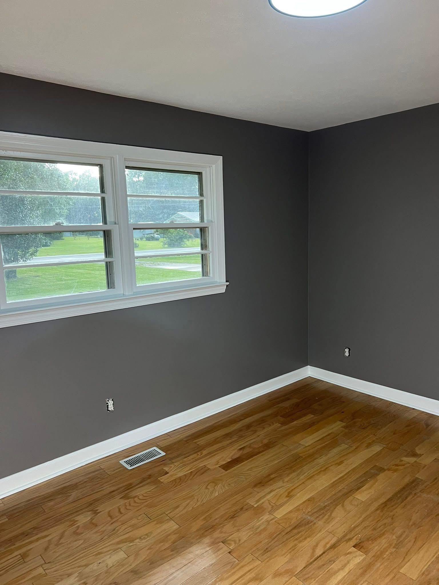 Empty room with gray walls, white trim, and hardwood floor. A window is on the left wall, ceiling light.