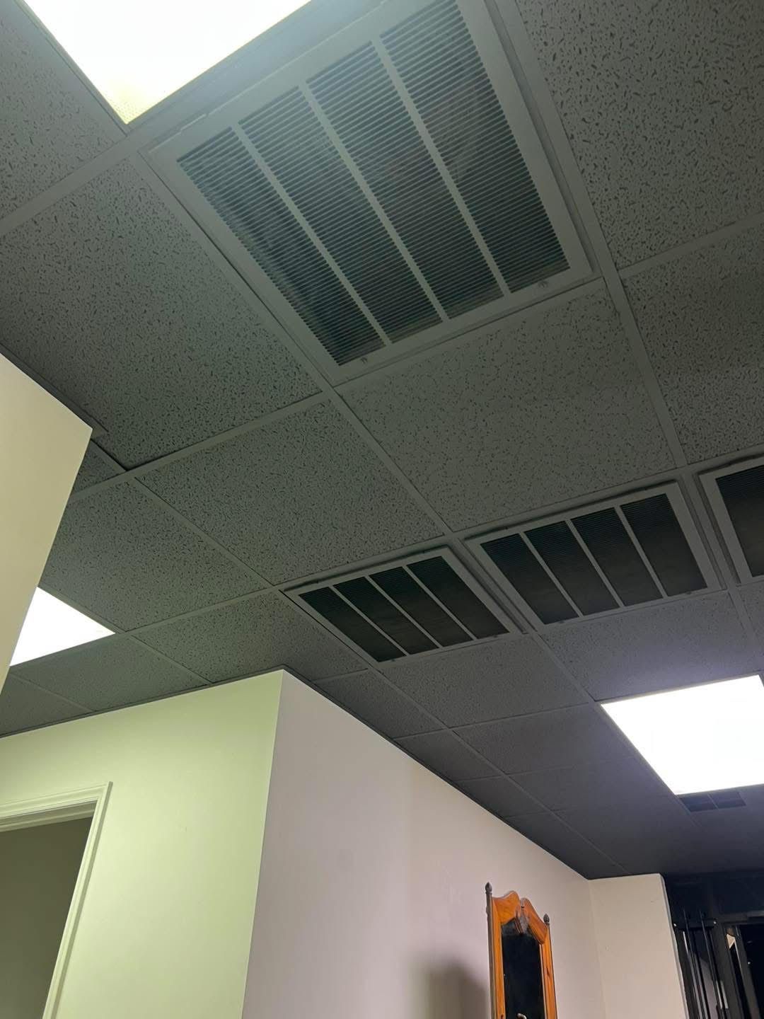 White ceiling tiles with air vents and recessed lights. Beige walls and doorway.