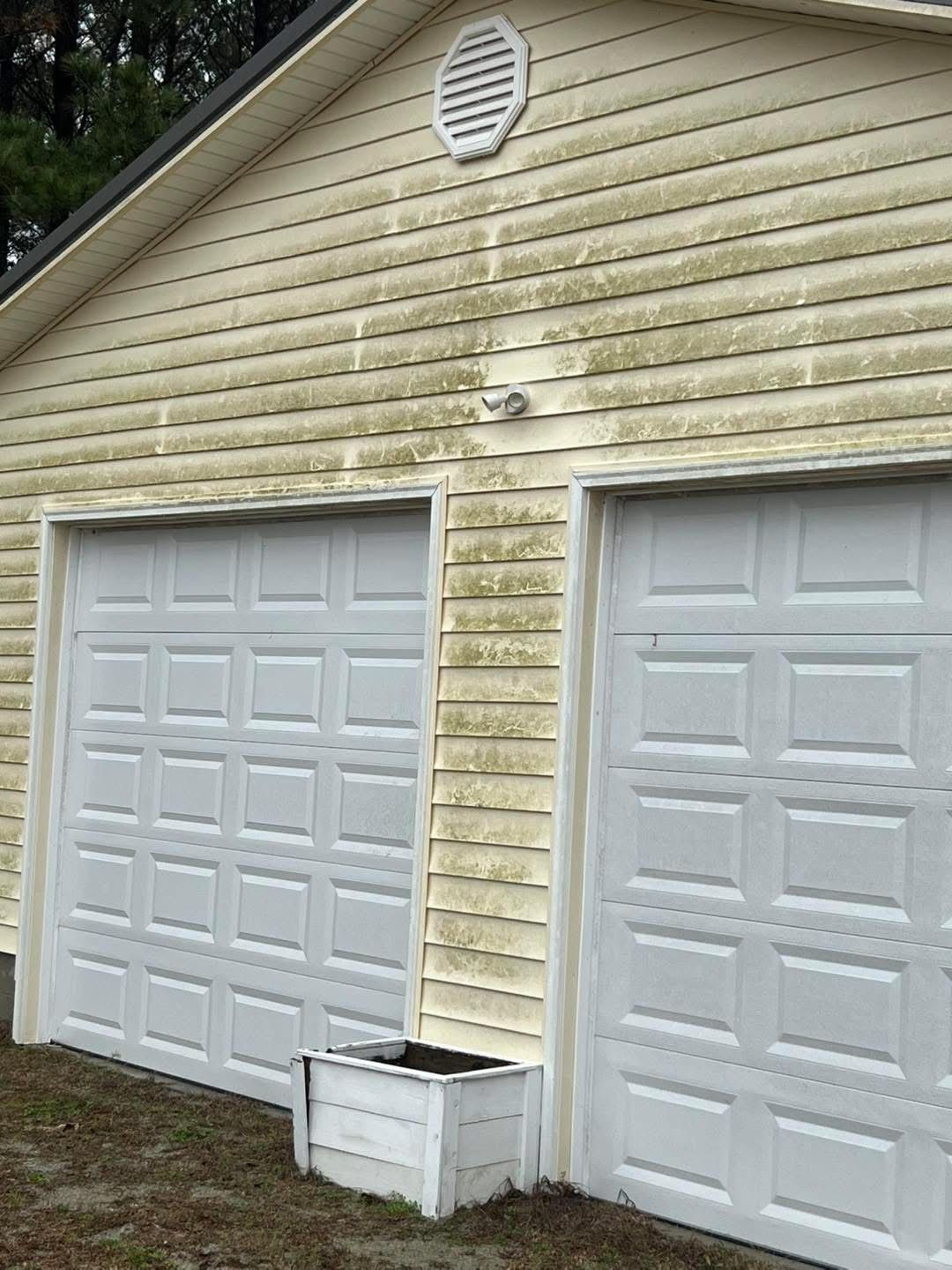 Yellow siding on a two-car garage covered with green algae, white garage doors, and a planter.