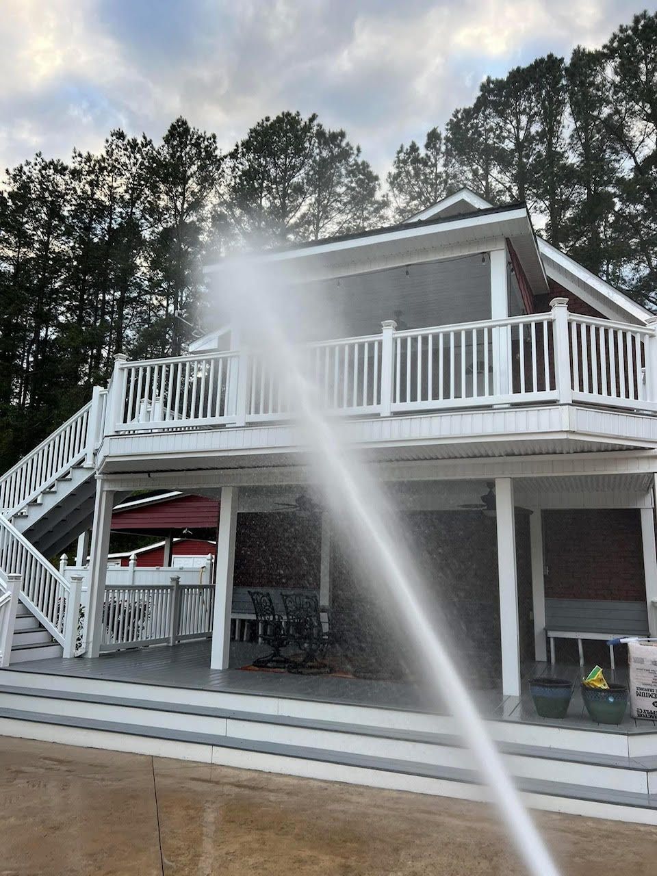 Pressure washing a two-story house with white trim, red accents, and a deck, near trees.