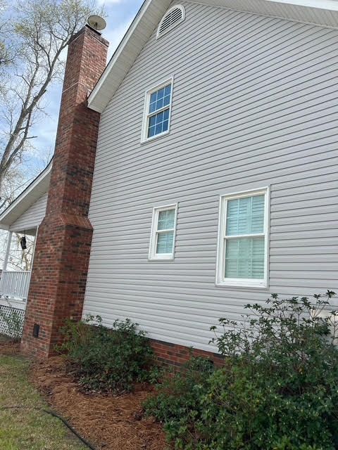 Side view of a house with gray siding, brick chimney, and white-framed windows, surrounded by bushes.
