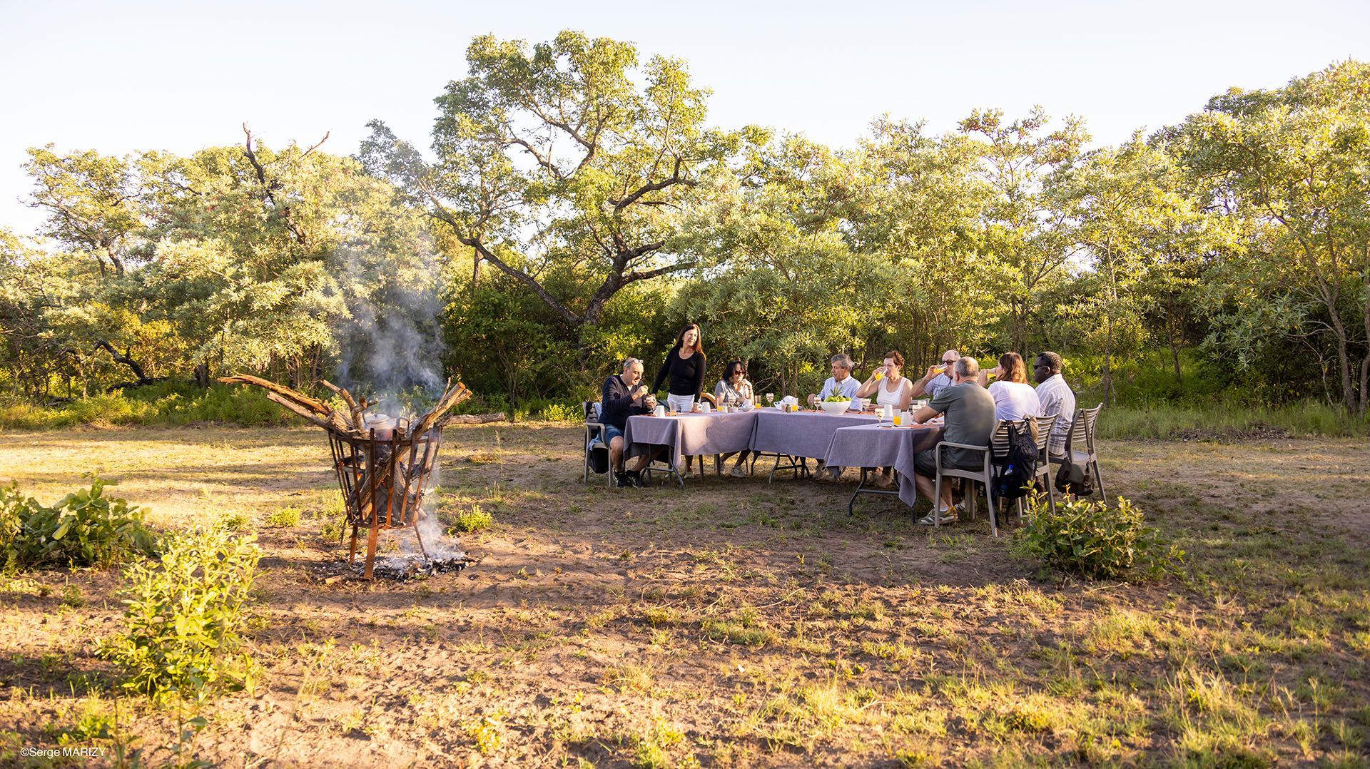 People seated at a long outdoor table in a sunny orchard or garden, with a smoking fire pit nearby