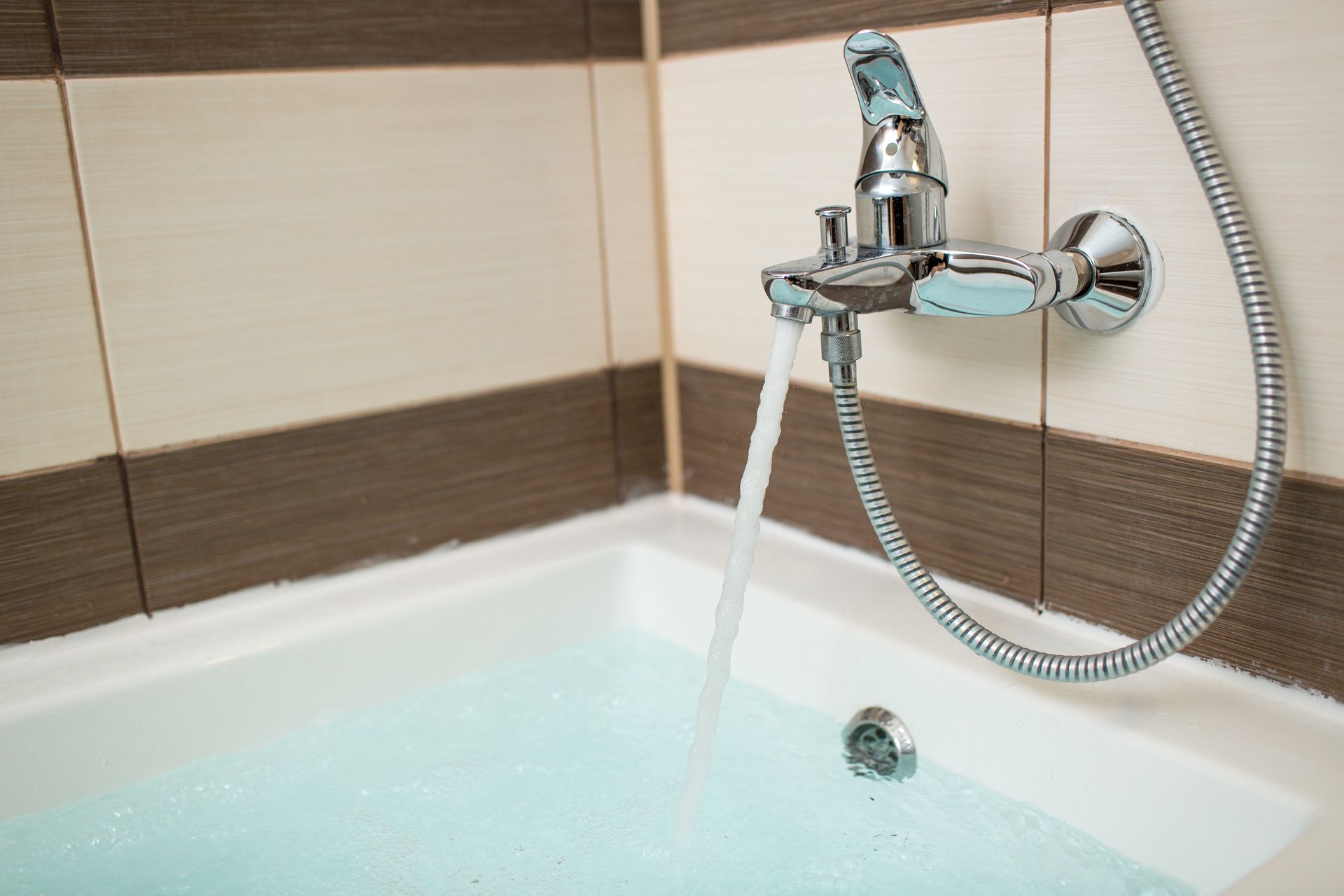 Water flows from a chrome bathroom faucet into a partially filled white bathtub against a tiled wall.