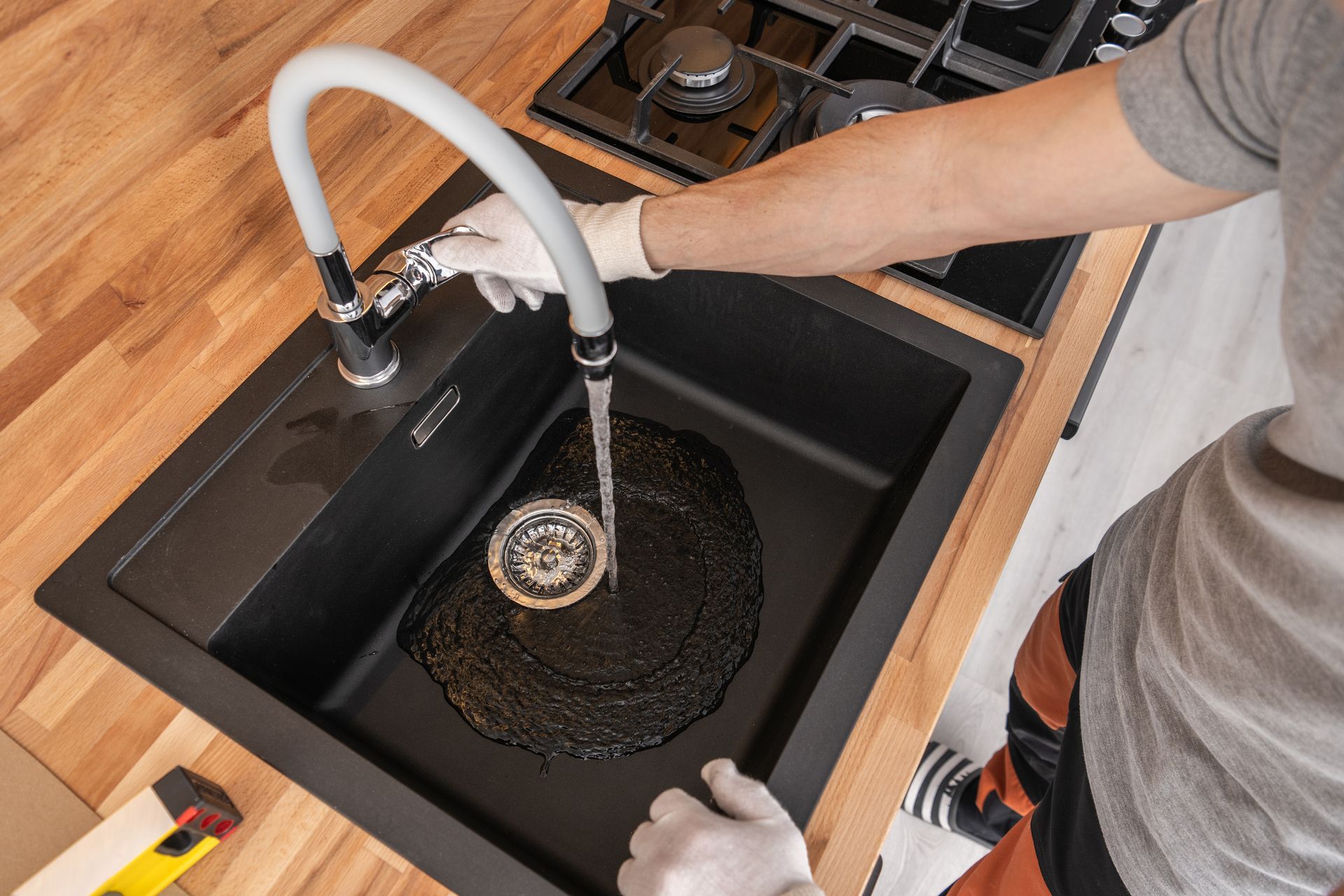 A gloved hand adjusts a white kitchen faucet, running water into a black sink in a room with a wooden countertop.