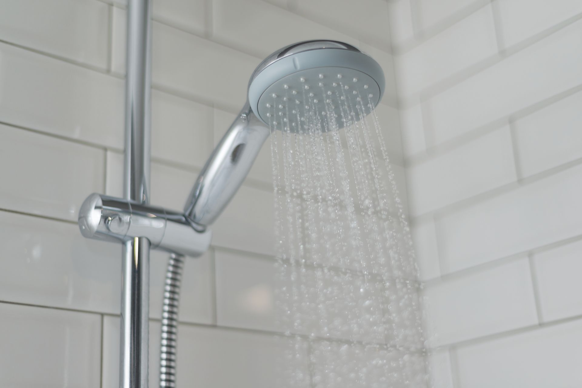 A chrome handheld showerhead mounted on a vertical bar, spraying water against white subway-style tiled walls.