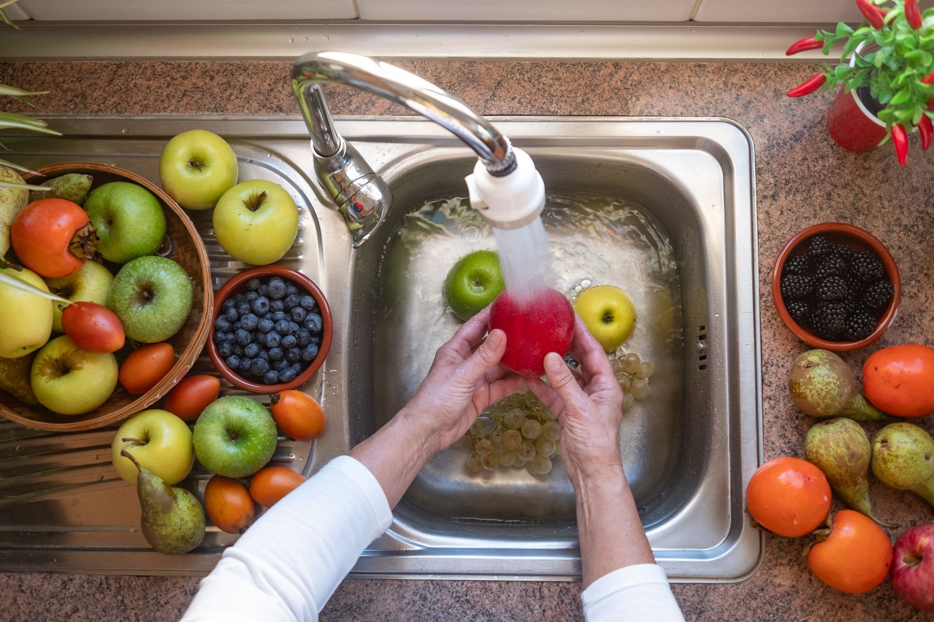 Hands wash a red apple under a kitchen sink faucet, surrounded by fresh fruits and berries on a marble countertop.
