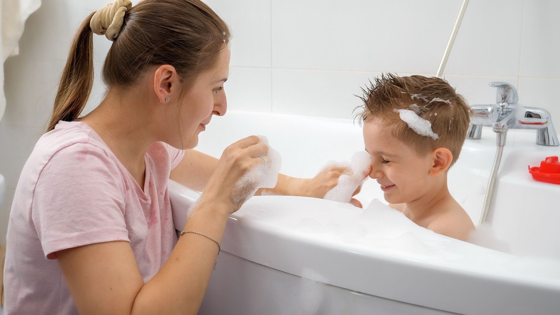A person in a light pink shirt bathes a child in a white tub, applying soap to their face amidst bubbles.