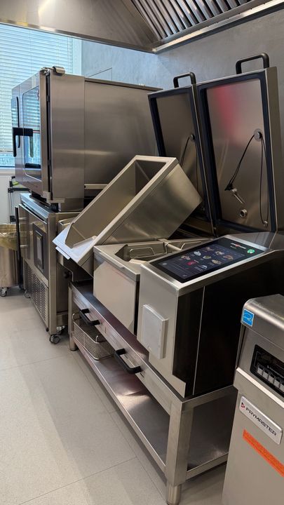 Attrezzature e contenitori da cucina in acciaio inossidabile allineati in un'area di preparazione commerciale con scaffalature metalliche.