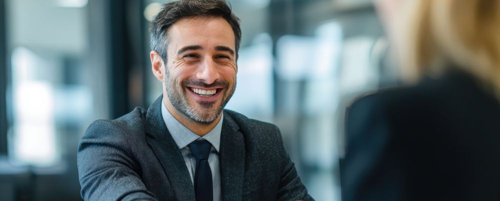 A smiling professional in a suit and tie during an office meeting.