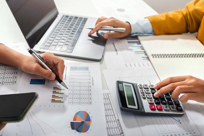 Two people analyzing financial charts and data on a desk with a laptop, calculator, and smartphone.