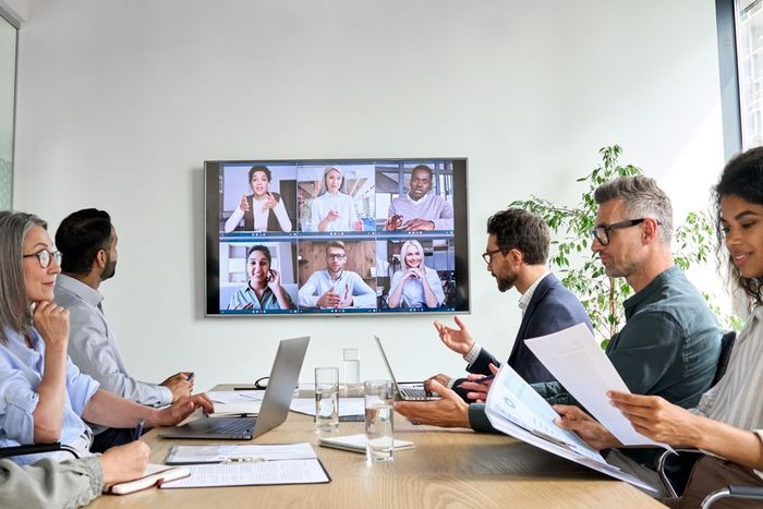 A diverse team in a conference room having a video conference call on a large screen mounted on the wall.
