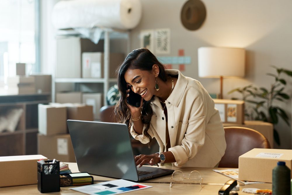 A smiling person in a light-colored shirt works on a laptop at a desk while talking on a phone in a home office.