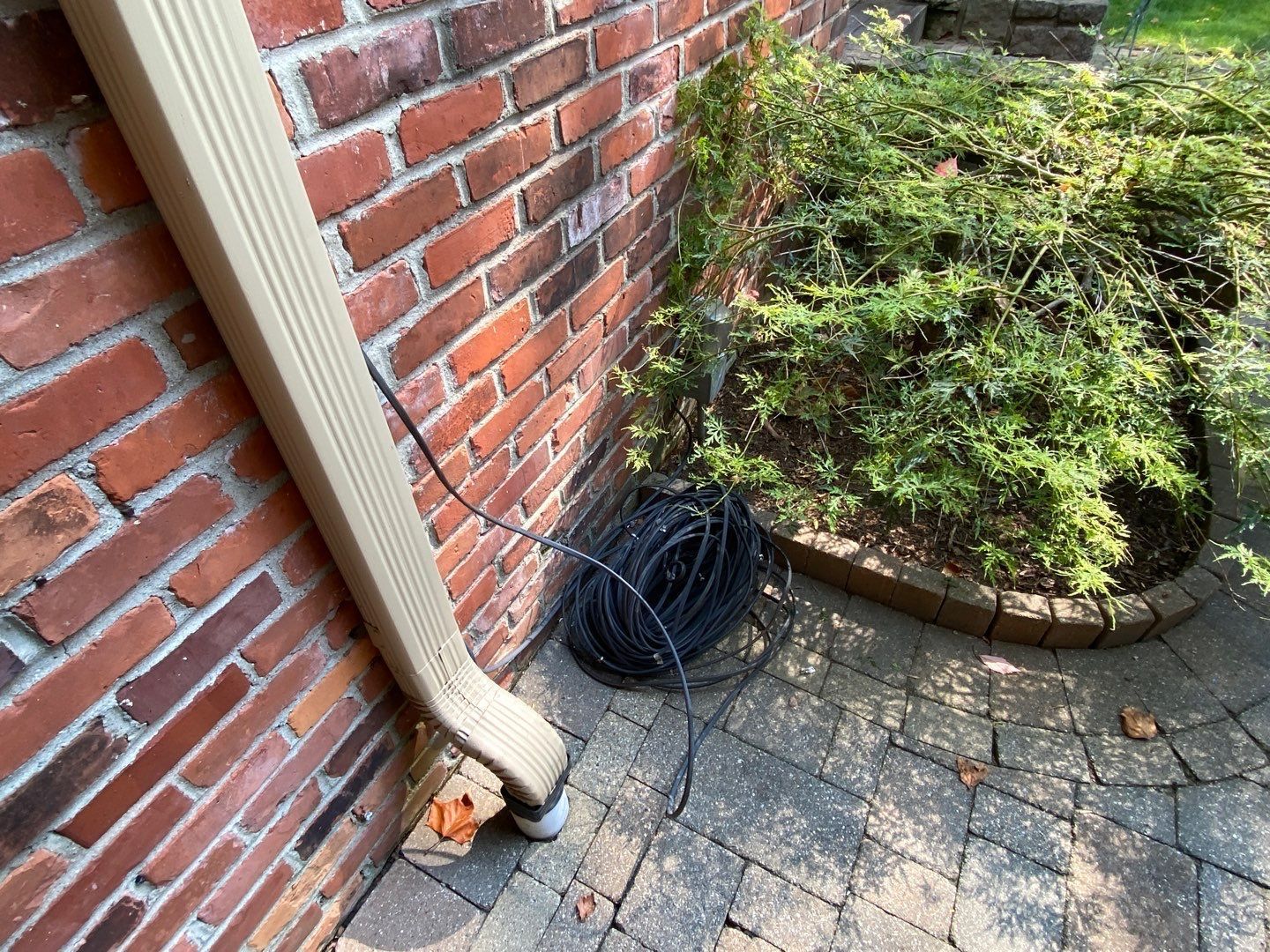 Brick house corner with downspout, coiled black cable, and curved garden bed beside a stone patio.
