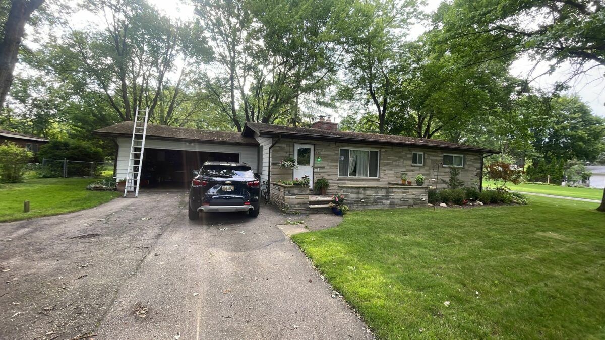 Single-story stone house with a car in the driveway and a ladder near the garage, surrounded by trees and lawn.